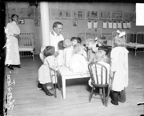 Portrait of Thornton W. Burgess, writer of the daily Bedtime Stories column in the Chicago Daily News, with children at the Home for Destitute Crippled Children. Burgess is wearing a surgical gown and is kneeling by a group of children sitting on a low table. Two of the children have crutches. A row of hospital beds and a nurse are visible in the background. This image was taken in Chicago, Illinois.